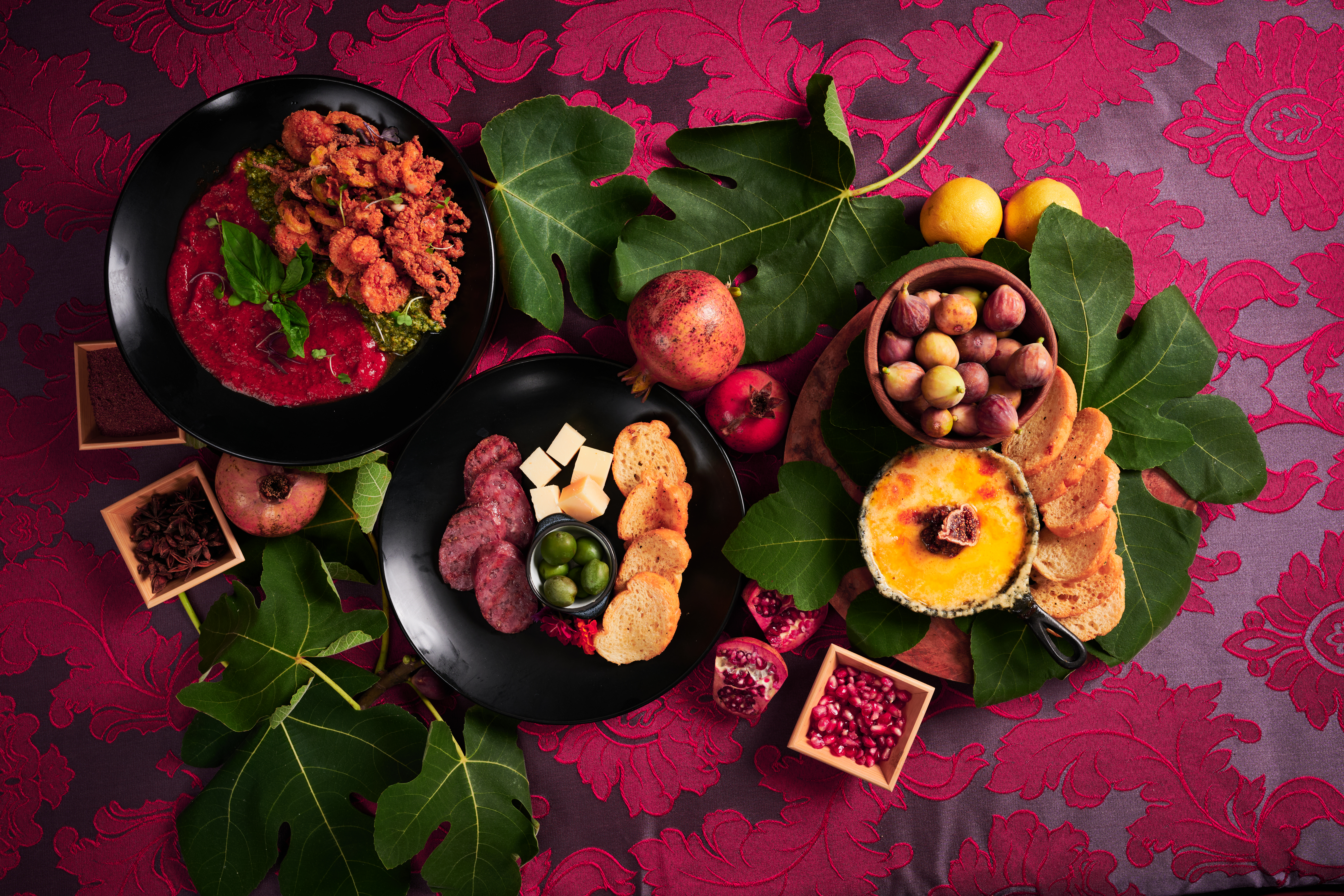 Variety of foods in dishes clustered with fruits and green leaves on a burgundy tablecloth